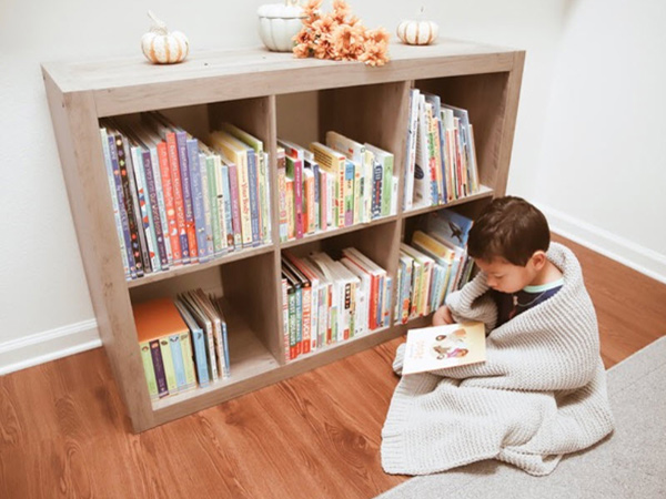 child wrapped in a blanket reading a picture book next to a bookshelf